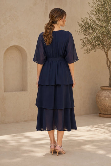 Navy blue tiered mother of the bride dress with modest closed back, sheer elbow-length sleeves, defined waist and flowing layered midi skirt, back view.