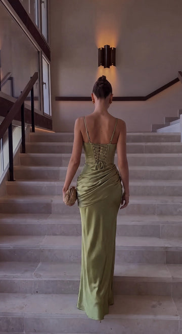 Back view of olive green prom dress featuring thin shoulder straps, lace-up corset back, crisscross ties, ruched hips, and flowing floor-length skirt.