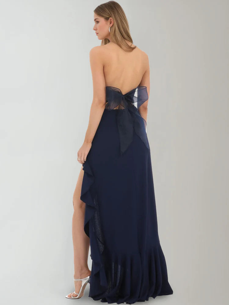 Back view of midnight navy strapless evening gown with open back, sheer bow detail, and ruffle slit skirt