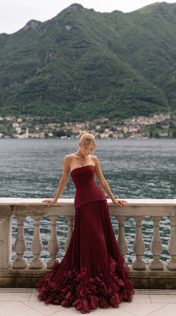 Vedress burgundy strapless wedding guest dress with a structured corset bodice, long pleated skirt and dimensional flower hem.