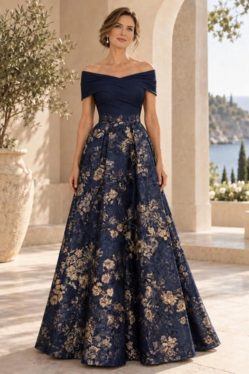 Navy blue A-line mother of the bride dress featuring off-shoulder ruched bodice, gold floral waistband and full floor-length floral patterned skirt