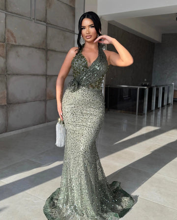 Sage green sequin mermaid prom dress featuring a halter V-neckline, asymmetric sparkling bodice, fitted waist and floor-length flared skirt.